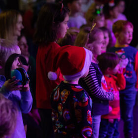 A crowd of yung children stood up in a dimly lit room watching something. Some of them have santa hats on.