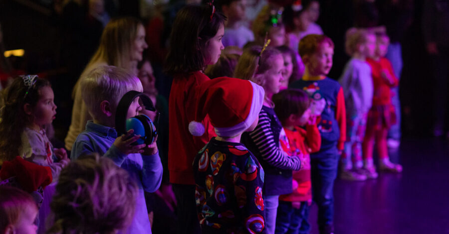 A crowd of yung children stood up in a dimly lit room watching something. Some of them have santa hats on.