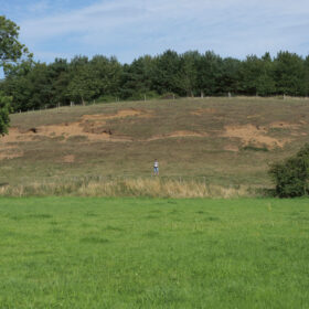 A view across a field with a hill in the background. A person stands in the distance.