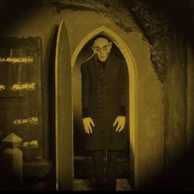 A black and white still of a man with distorted features walking through an arched doorway