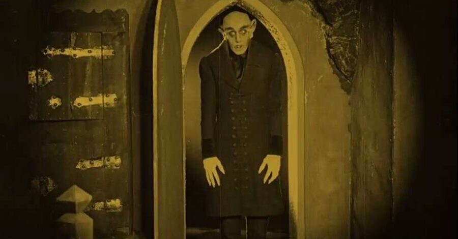 A black and white still of a man with distorted features walking through an arched doorway