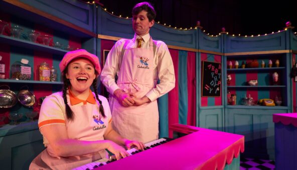 Two performers wearing pink ice-cream shop uniforms. One is sat down playing a piano. The stage setting looks like a shop.
