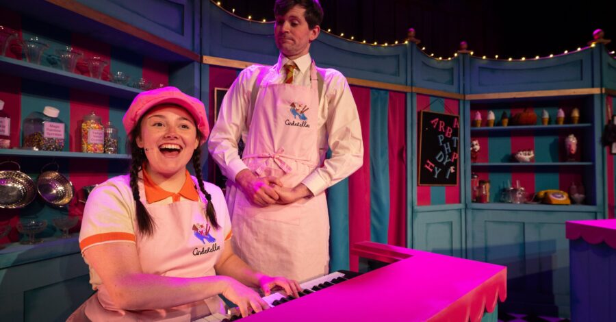 Two performers wearing pink ice-cream shop uniforms. One is sat down playing a piano. The stage setting looks like a shop.
