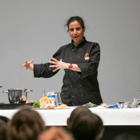 A female chef in a black coat demonstrates cooking techniques on a stage, surrounded by pots, pans, and ingredients, engaging with an audience.