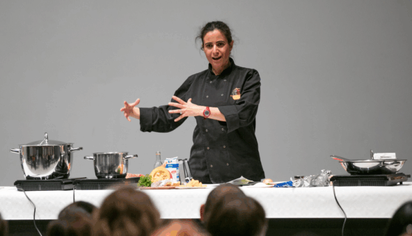 A female chef in a black coat demonstrates cooking techniques on a stage, surrounded by pots, pans, and ingredients, engaging with an audience.
