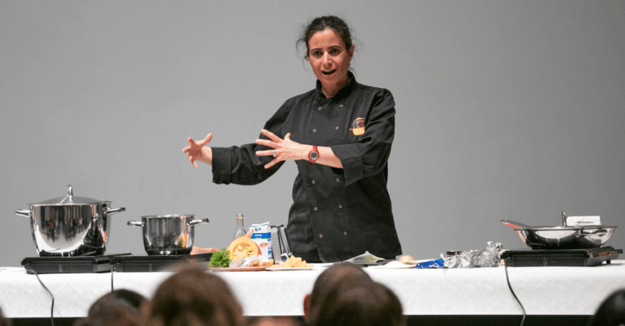 A female chef in a black coat demonstrates cooking techniques on a stage, surrounded by pots, pans, and ingredients, engaging with an audience.