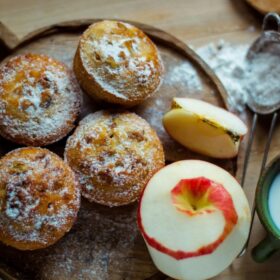 Five golden-brown muffins dusted with powdered sugar are arranged on a rustic wooden cutting board. Two fresh apples are next to them: one whole apple, mostly peeled with the skin spiraling around it, and a slice of apple. To the right, a dark green mug holds a small amount of milk or cream, and a small metal sifter with powdered sugar sits partially behind the apple slice. The scene is warmly lit.