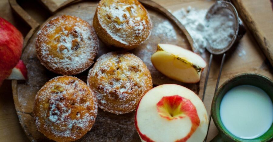Five golden-brown muffins dusted with powdered sugar are arranged on a rustic wooden cutting board. Two fresh apples are next to them: one whole apple, mostly peeled with the skin spiraling around it, and a slice of apple. To the right, a dark green mug holds a small amount of milk or cream, and a small metal sifter with powdered sugar sits partially behind the apple slice. The scene is warmly lit.