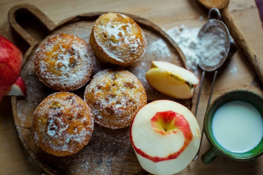 Five golden-brown muffins dusted with powdered sugar are arranged on a rustic wooden cutting board. Two fresh apples are next to them: one whole apple, mostly peeled with the skin spiraling around it, and a slice of apple. To the right, a dark green mug holds a small amount of milk or cream, and a small metal sifter with powdered sugar sits partially behind the apple slice. The scene is warmly lit.