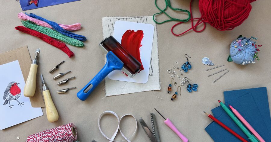 A table with a range of crafting materials and tools on it. There is green and red wool, string, scissors, crafting tools, and a range of different coloured paper and card.