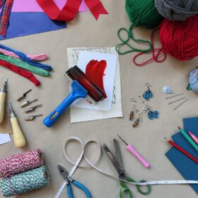 A table with a range of crafting materials and tools on it. There is green and red wool, string, scissors, crafting tools, and a range of different coloured paper and card.