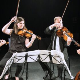 a string quartet dressed in black and playing in front of a black cloth, two have their bows dramatically in the air