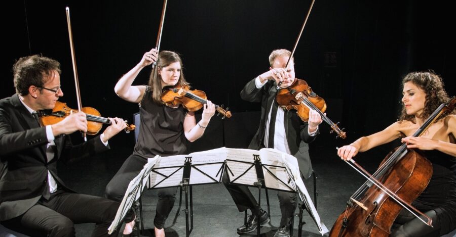 a string quartet dressed in black and playing in front of a black cloth, two have their bows dramatically in the air