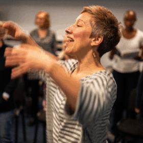 A woman with short grey cropped hair wearing a grey and white striped top stretches her arms out as if she is conducting a choir.