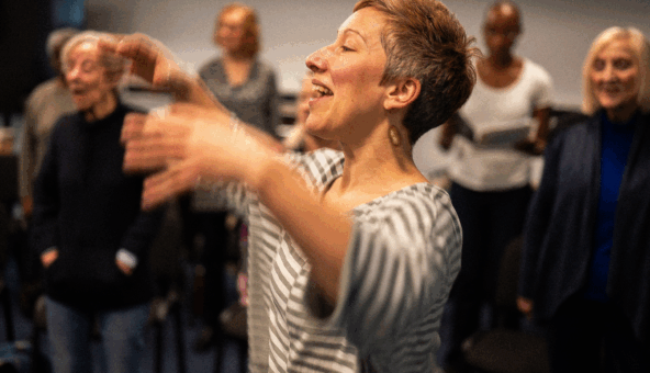 A woman with short grey cropped hair wearing a grey and white striped top stretches her arms out as if she is conducting a choir.