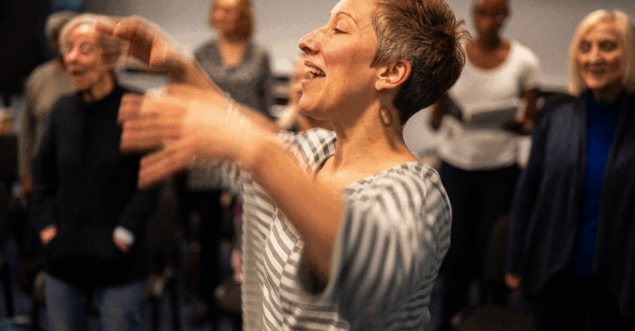 A woman with short grey cropped hair wearing a grey and white striped top stretches her arms out as if she is conducting a choir.