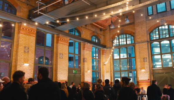 Inside the Engine Hall at the People's History Museum in the evening. There are people stood about and warm white festoon lighting hanging.