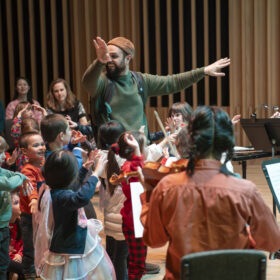 A performer stamds with his arms raised up at his sides. There are children stood around him, watching him and raising their arms up too.