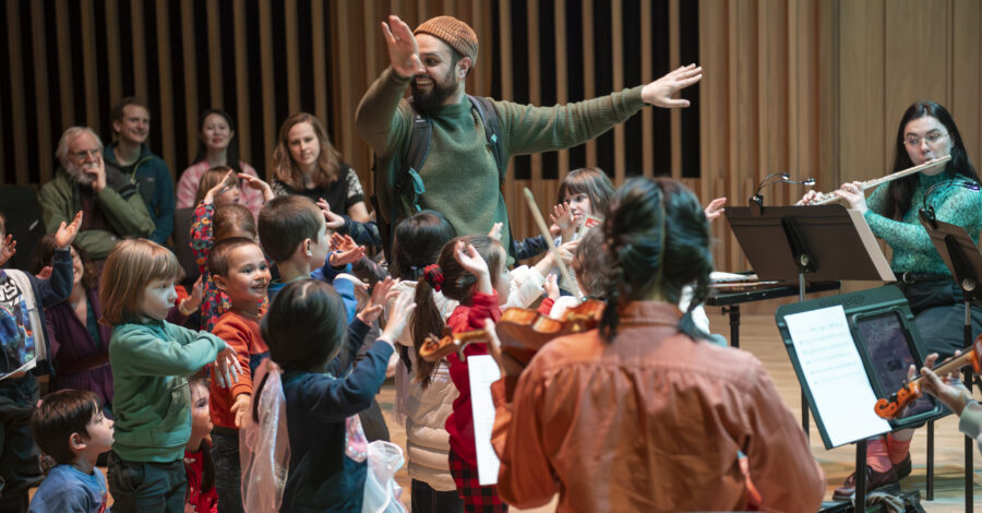 A performer stamds with his arms raised up at his sides. There are children stood around him, watching him and raising their arms up too.