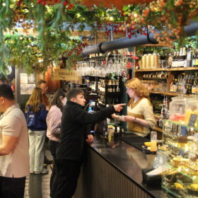 People standing at a counter in a cafe bar. There are drinks and cakes on display.