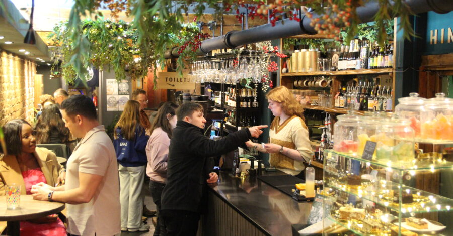People standing at a counter in a cafe bar. There are drinks and cakes on display.