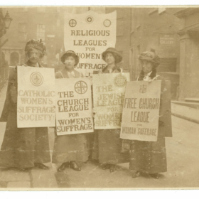 A black and white image of four women holding placards that read 'The Church league of Women's Suffrage', 'Catholic Women's Suffrage Society' and 'Free Church League for Women's Suffrage'.