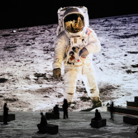 A large projection of an astronaut standing on the moon. Visitors to the exhibition look tiny incomparison and sit, move and stand as they experience the exhibition.