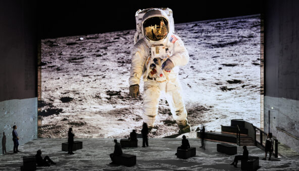 A large projection of an astronaut standing on the moon. Visitors to the exhibition look tiny incomparison and sit, move and stand as they experience the exhibition.