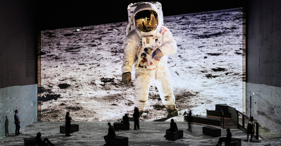 A large projection of an astronaut standing on the moon. Visitors to the exhibition look tiny incomparison and sit, move and stand as they experience the exhibition.