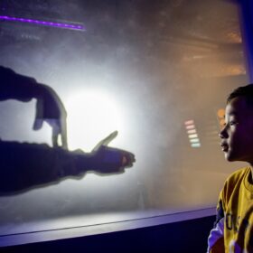 A young boy, wearing a yellow sweatshirt, looking at a pair of hands behind a shadow wall.