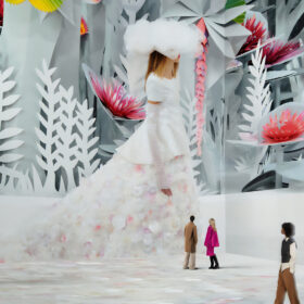 A large-scale video installation of a model walking down a cat walk. She is wearing a long white gown with a train and a large white feathered hat. There a re four people experiencing the artwork - they look tiny because the video installation is on such a large screen.