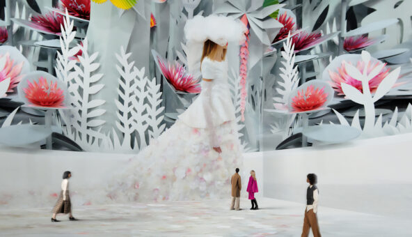 A large-scale video installation of a model walking down a cat walk. She is wearing a long white gown with a train and a large white feathered hat. There a re four people experiencing the artwork - they look tiny because the video installation is on such a large screen.