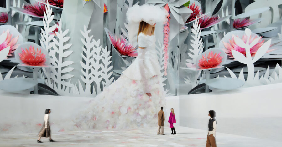 A large-scale video installation of a model walking down a cat walk. She is wearing a long white gown with a train and a large white feathered hat. There a re four people experiencing the artwork - they look tiny because the video installation is on such a large screen.