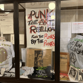 A display cabinet from Manchester Central Library's current exhibition: Punk: The Art of Rebellion. The display houses a banner and two t-shirts with slogans from the punk era.