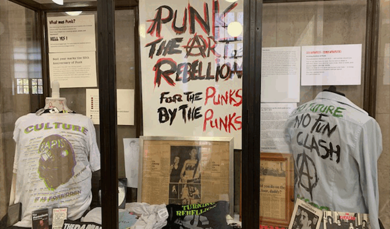 A display cabinet from Manchester Central Library's current exhibition: Punk: The Art of Rebellion. The display houses a banner and two t-shirts with slogans from the punk era.