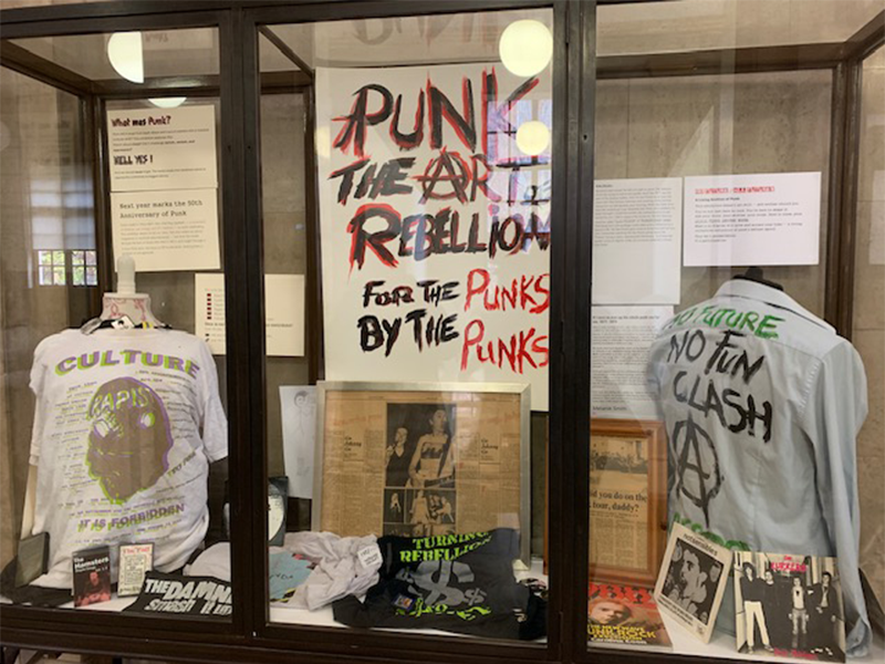 A display cabinet from Manchester Central Library's current exhibition: Punk: The Art of Rebellion. The display houses a banner and two t-shirts with slogans from the punk era.