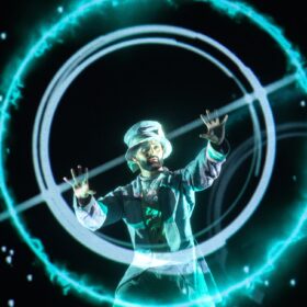 A performer wearing a silver top hat and suit with his arms outstretched, as if he is casting a spell.