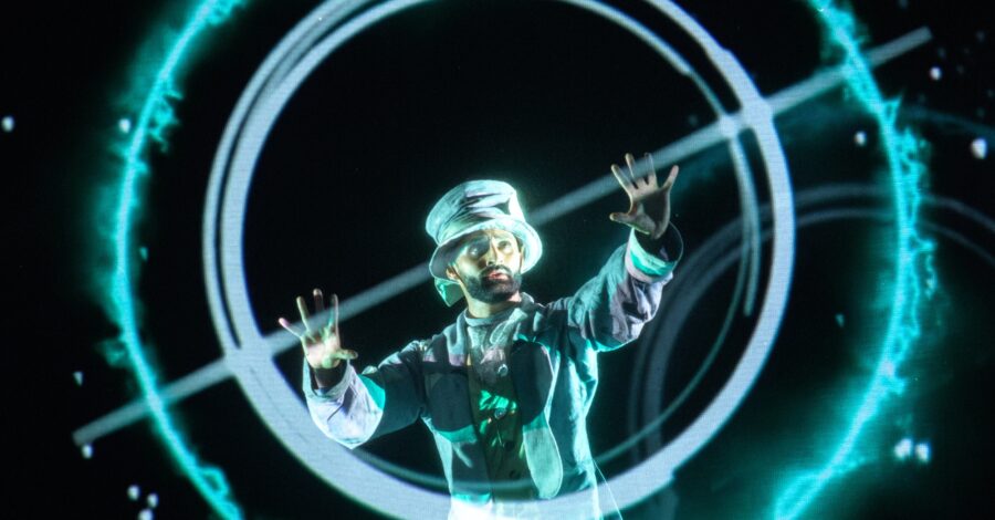 A performer wearing a silver top hat and suit with his arms outstretched, as if he is casting a spell.