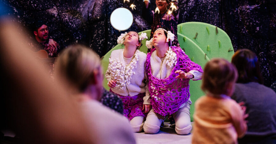 Performers in white and purple costumes kneel on stage, surrounded by a colourful, nature-themed set, as children watch the show in the audience.