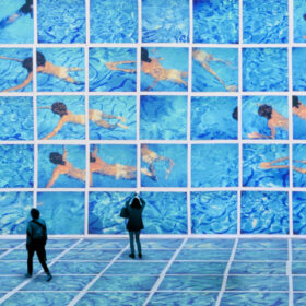 A huge video installation of David Hockney's artwork. It looks like a swimming pool with several people swimming in it and two visitor stand looking at it.