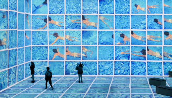 A huge video installation of David Hockney's artwork. It looks like a swimming pool with several people swimming in it and two visitor stand looking at it.