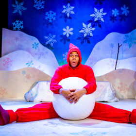 A performer in a bright red costume sits on a snowy stage set, holding a large snowball between their legs with a surprised expression. The colourful winter backdrop features snowflakes, hills, a snowman, and a traffic light with glowing lights.
