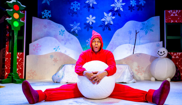 A performer in a bright red costume sits on a snowy stage set, holding a large snowball between their legs with a surprised expression. The colourful winter backdrop features snowflakes, hills, a snowman, and a traffic light with glowing lights.