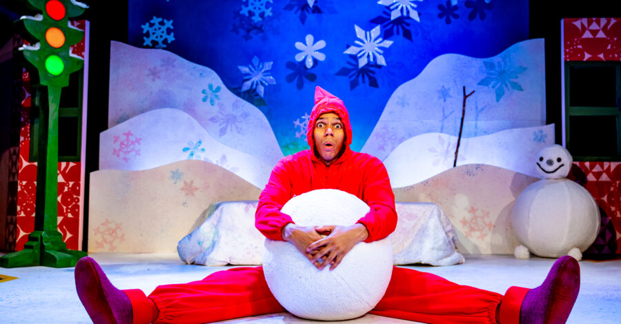 A performer in a bright red costume sits on a snowy stage set, holding a large snowball between their legs with a surprised expression. The colourful winter backdrop features snowflakes, hills, a snowman, and a traffic light with glowing lights.