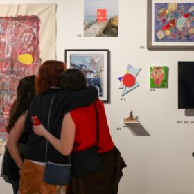 Three people with their arms around each other looking at artwork on a gallery wall.