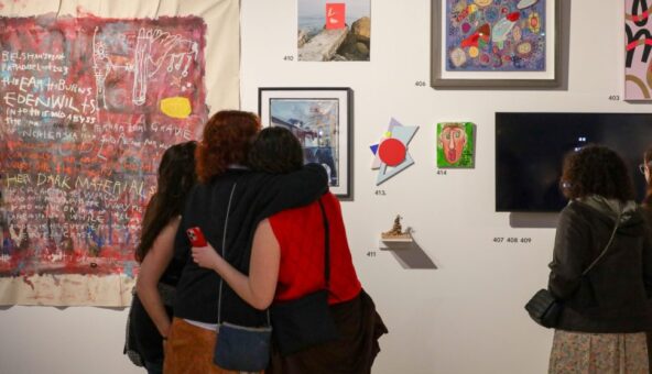 Three people with their arms around each other looking at artwork on a gallery wall.