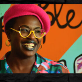 Artist Jasmin Issaka is smiling, and wears a red hat, yellow-framed glasses and a brightly coloured shirt.