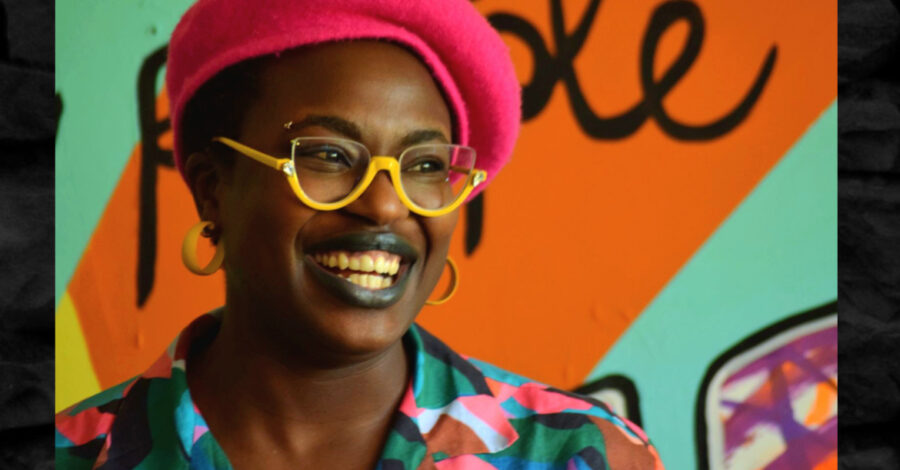 Artist Jasmin Issaka is smiling, and wears a red hat, yellow-framed glasses and a brightly coloured shirt.