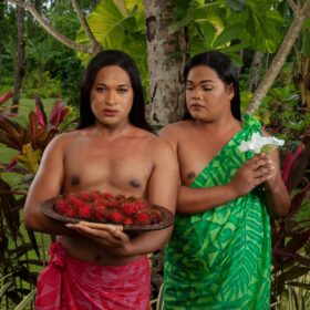Two people stood next to one another wearing traditional Pacific dress. One person wears a red sarong and holds a plate of red fruit and the other wears a green dress and holds a white flower.