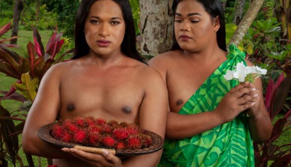 Two people stood next to one another wearing traditional Pacific dress. One person wears a red sarong and holds a plate of red fruit and the other wears a green dress and holds a white flower.